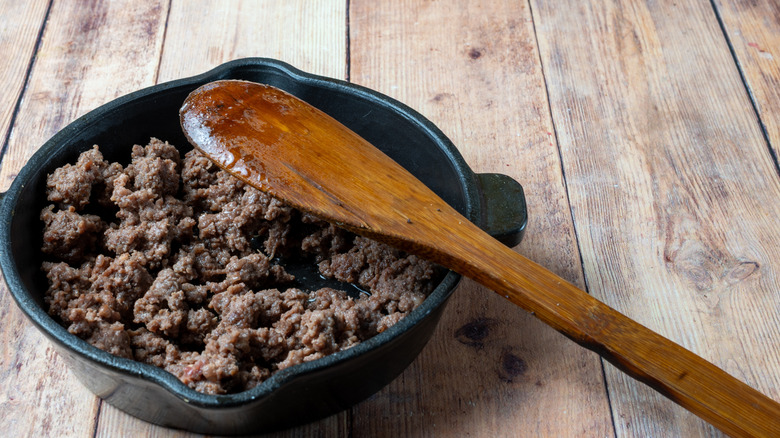 A cast iron skillet full of browned ground beef sitting on a wooden table with a wooden spoon resting over the edge of the skillet