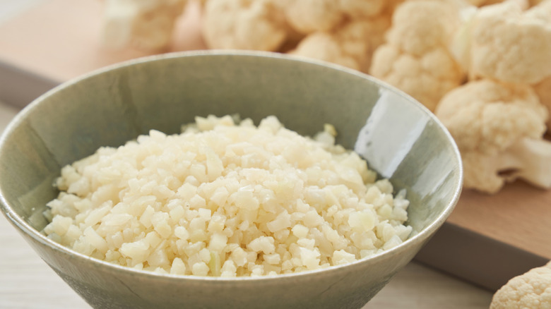 A bowl of rice cauliflower with cauliflower florets on a cutting board behind it
