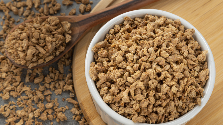 A bowl of textured vegetable protein sitting on a wooden cutting board with more scattered on the table and a spoon next to it