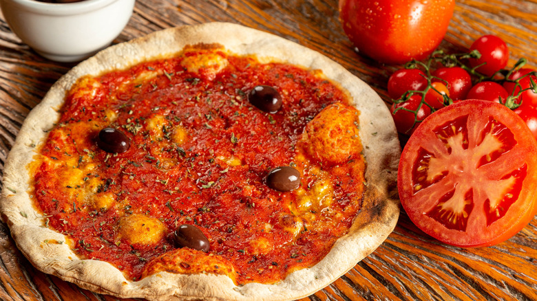 A pizza marinara topped with olives next to a cut tomato