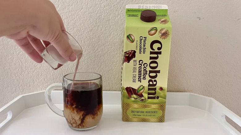 A person pouring creamer into a glass cup of coffee with a carton of Chobani Pistachio Chocolate Coffee Creamer nearby