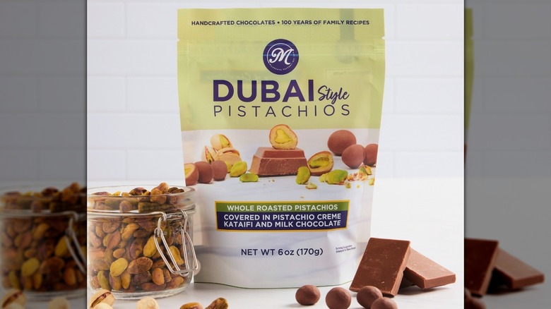 A bag of MilkShake Factory Dubai-Style Pistachios with a glass jar of pistachios and chocolate squares surrounding