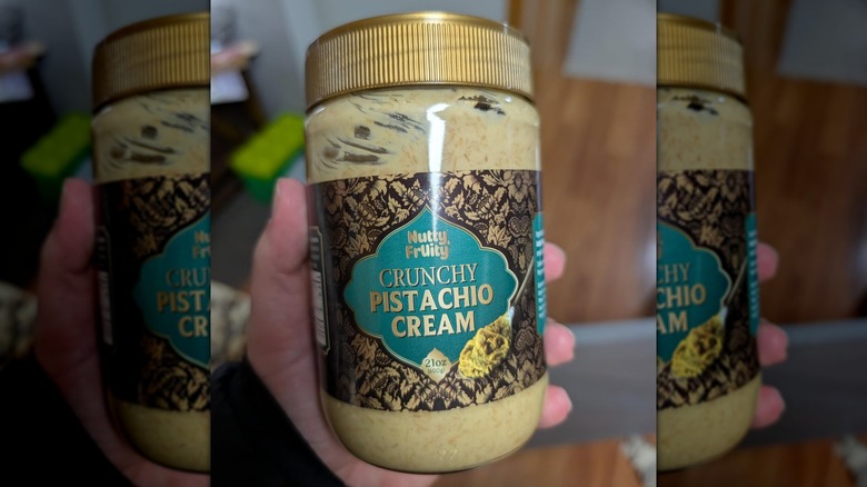 A person holding a small jar of partially eaten Nutty & Fruity Crunchy Pistachio Cream