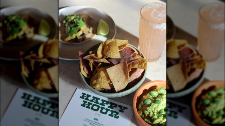 Happy hour menu on the table with chips, guacamole, a taco, and a cocktail