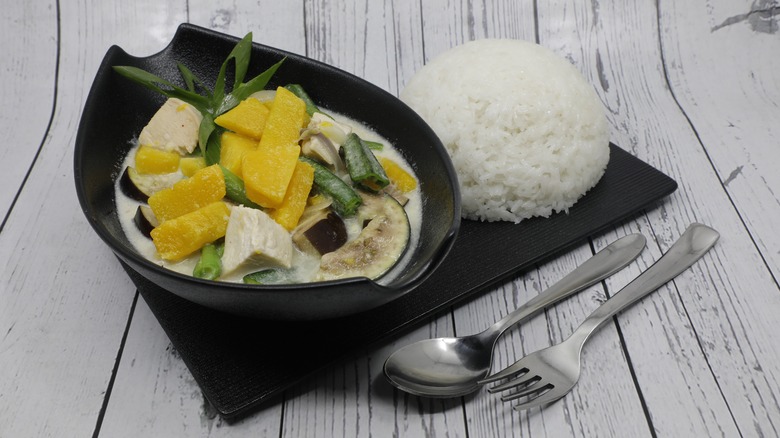 A black bowl filled with vegetables in a creamy sauce alongside a scoop of white rice on a white wooden table.