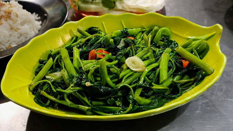 A leaf-shaped dish of stir-fried kangkong leaves with garlic, served on a metal countertop along with a bowl of rice.