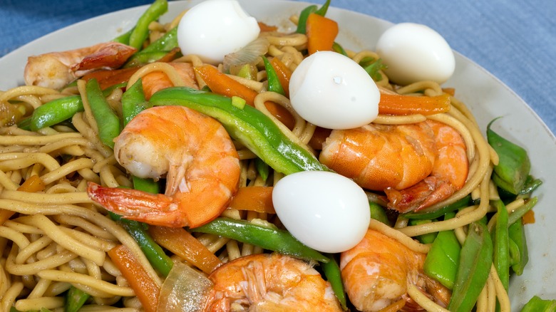A dish of thick lo mein noodles with shrimp, quail eggs, and snap peas on a light blue tablecloth.