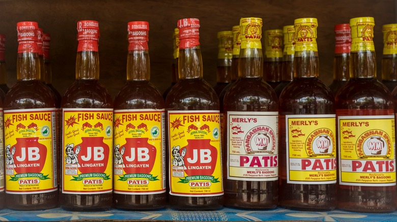 Rows of bottles of patis fish sauce on a shelf. The bottles all have yellow, white, and red labels depicting jars alongside various brand names and logos, along with red or yellow plastic-wrapped caps.