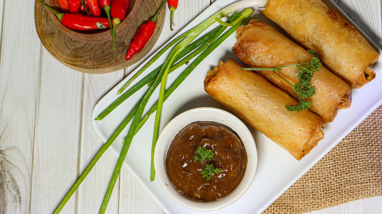 A plate of three fried lumpia spring rolls with a reddish-brown dipping sauce on a white wooden table.