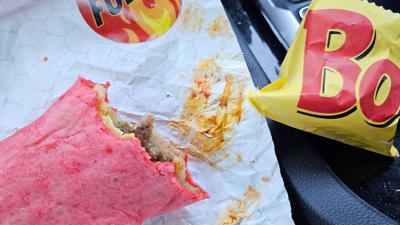 Apartially eaten burrito with a red tortilla sitting on a piece of paper next to a wrapper that says "Bo"