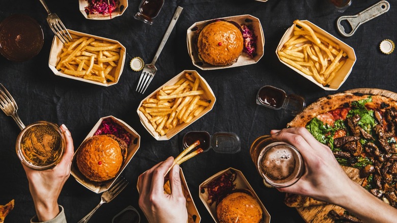 A selection of fast food items including burgers, fries, and a pizza, with people's hands holding drinks