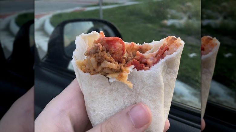 A hand holding a partially eaten Beefy Crunch Burrito in front of a car window