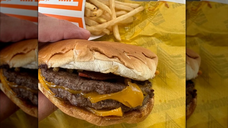 A hand holding a Whataburger double cheeseburger over some paper with fries in the background