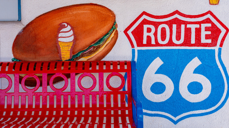 Wall of a fast food restaurant with art of a burger, ice cream, and Route 66 sign, with a red bench in front of it