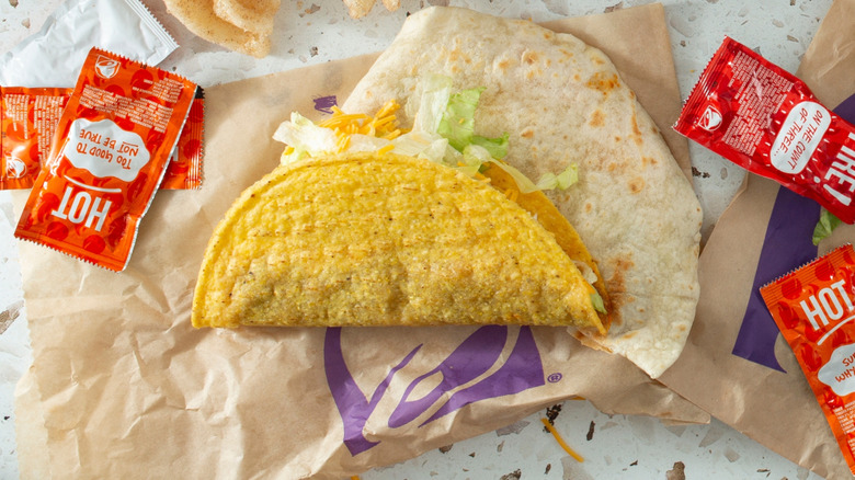 Taco Bell Crunchy Taco resting atop a tortilla and paper bag and surrounded by hot sauce packets