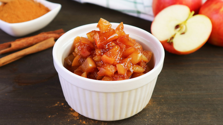 Caramelized apples in white bowl next to cinnamon sticks and fresh apples
