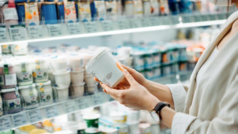 Person reading the ingredients list on a yogurt container in a store aisle