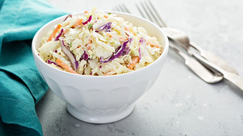 coleslaw in a white bowl next to knife and fork and blue tablecloth