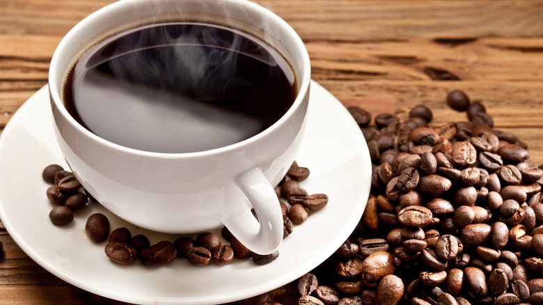 Cup of hot black coffee with steam coming out on wooden table with roasted coffee beans