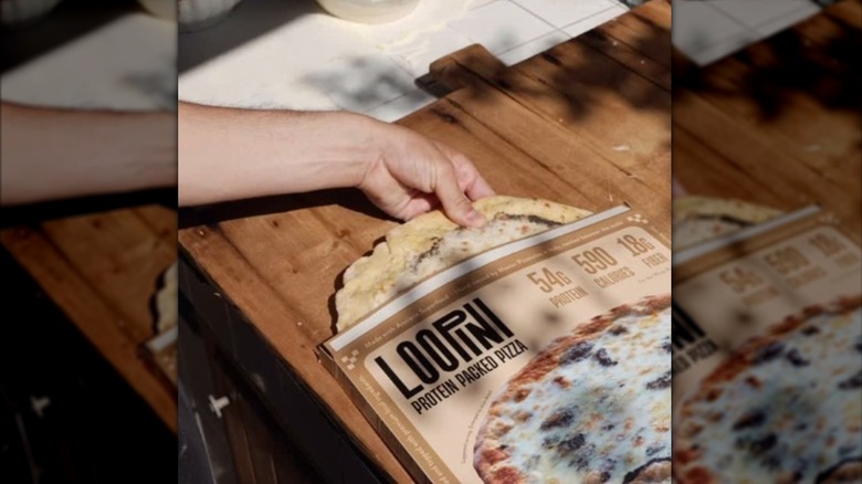 A hand removie a Loopini protein pizza from the box on a wooden counter