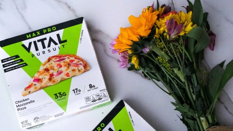 A box of Vital Pursuit Max Pro Chicken Mozzarella Pizza sitting next to some flowers on a counter