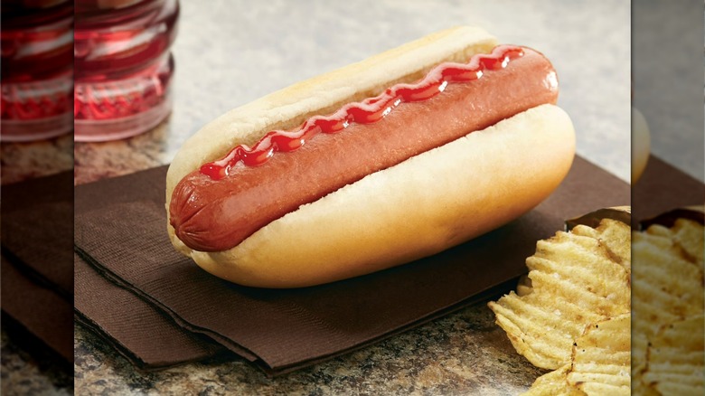 Ball Park frank topped with ketchup next to potato chips and a glass