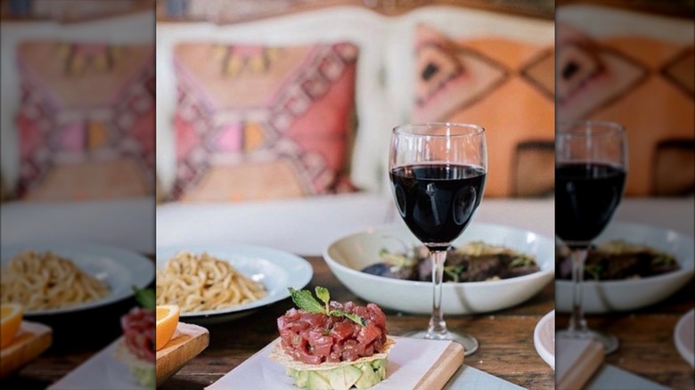 Tuna tartare, pasta, and wine on a rustic wood table with a colorful couch in the background