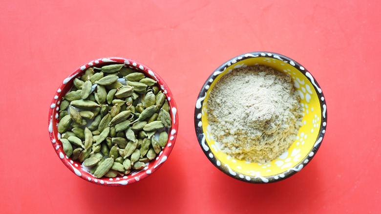 A red and white ceramic bowl filled with whole green cardamom pods and a black and white ceramic bowl with a yellow interior filled with ground cardamom, on a red surface
