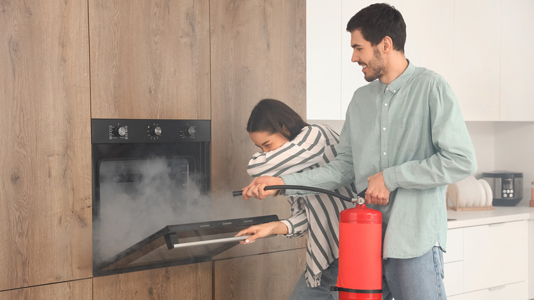A young man and women holding a red fire extinguisher putting out a fire in the oven, in a home kitchen