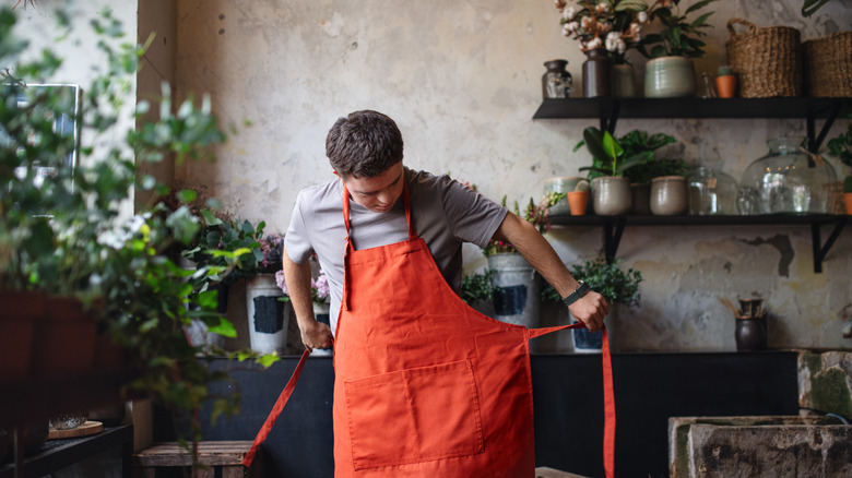 Person putting on a bright orange apron, standing in a ktichen filled with plants