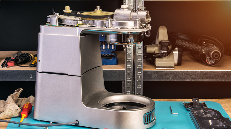 disassembled stand mixer on a workbench, surrounded by tools and parts, and with other items awaiting repair visible on a shelf to the rear