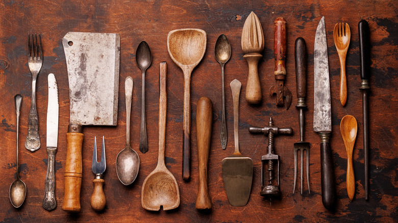 a selection of vintage kitchen utensils and silverware, including silver and wooden spoons, a fruit reamer, a corkscrew, an old-school can opener, and several knives and serving pieces