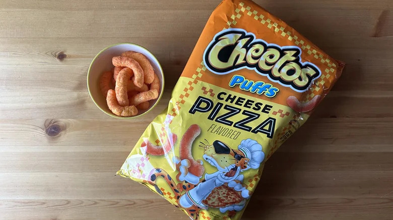 A bag of Cheetos Puffs Cheese Pizza Flavored on a table next to a small white bowl of puffs