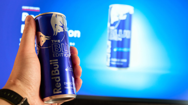 A close-up of a man holding a can of Red Bull Blue Edition in front of a television advertisement for the drink