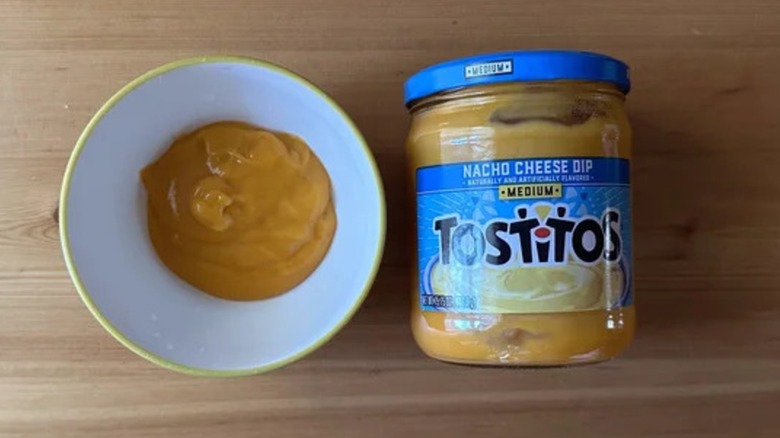 A jar of Tostitos Nacho Cheese Dip on a table next to a white bowl of the dip
