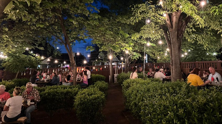 People at tables in beer garden with greenery, shrubs, and trees