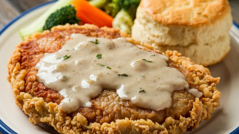 A close-up of chicken-fried steak with creamy gravy, a bsicuit, and veggies