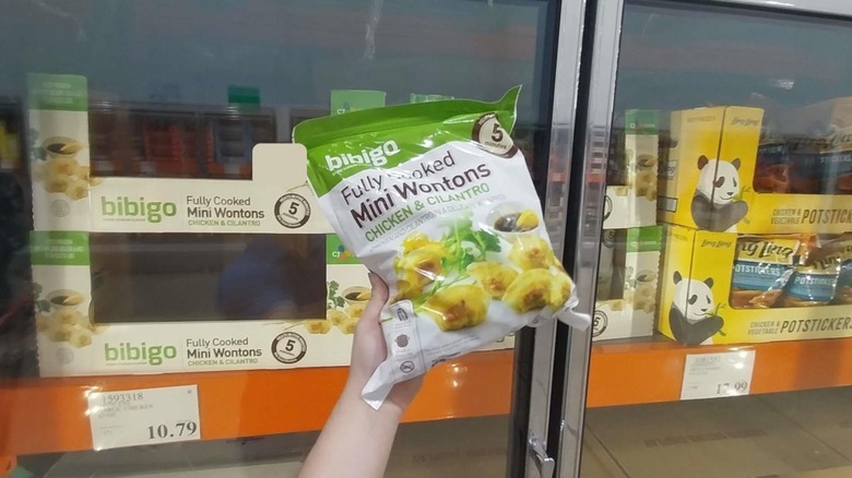 A hand holding up a bag of Bibigo Chicken & Cilantro Mini Wontons in front of a freezer in a Costco warehouse