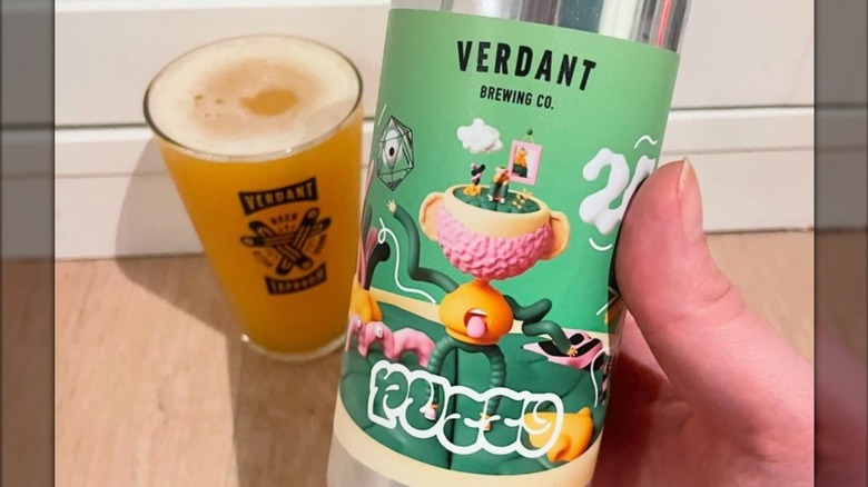 Person holding a green can of Verdant Brewing Co. Putty beer with a full glass in the background.