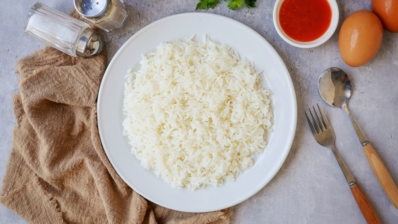 Top view of a plate of cooked white rice surrounded by eggs, utensils, and shakers