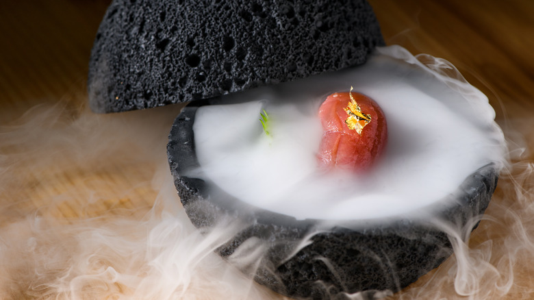 Gourmet dish in a dark bowl with smoke coming out and revealing a sushi roll with a gold leaf