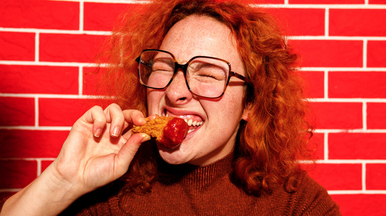 Woman in front of red brick background winces as she eats a saucy chicken wing