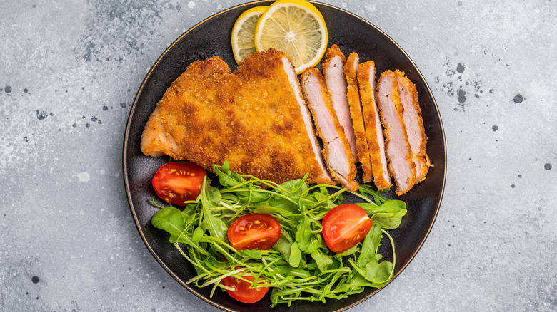 A piece of breaded veal whole and sliced served with salad and lemon slices on a dark plate