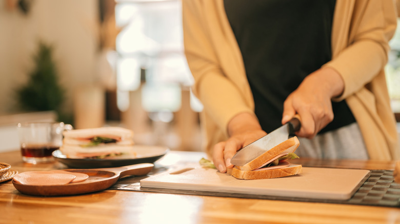 Close-up of a person in a brown cardigan and black tee is cutting a sandwich on a cutting board on a counter, sliced meat is at the side along with another sandwich