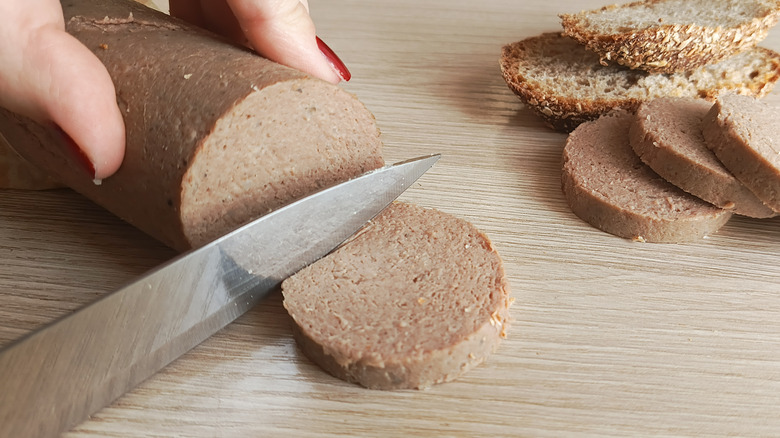 The close-up of liverwurst being sliced for a sandwich, the bread is in the background with other slices, on a countertop