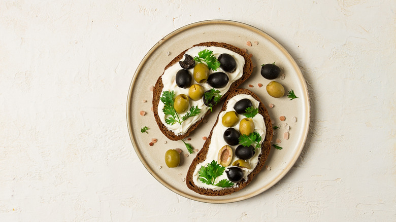 A cream cheese and black and green olive open-faced sandwich on dark bread, neutral plate, and neutral background