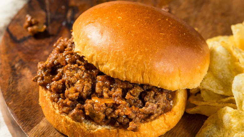 A close-up of a Sloppy Joe on a golden brown bun, beside plain potato chips, on a wooden cutting board