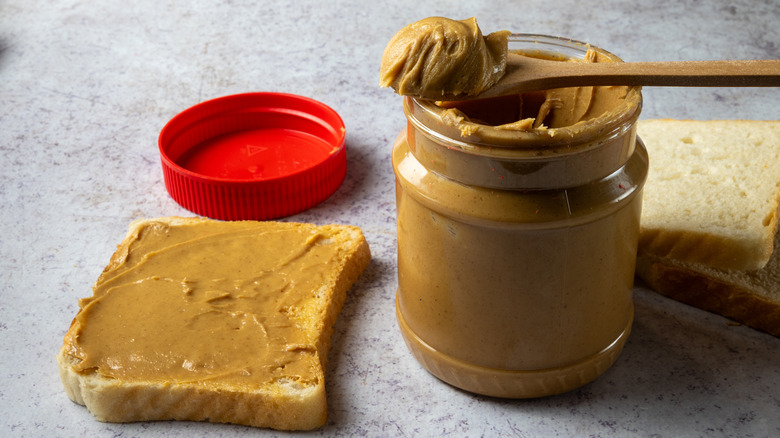 A jar of peanut butter, lid off with a spoonful balanced on top, already spread on one slice of bread with more behind the jar