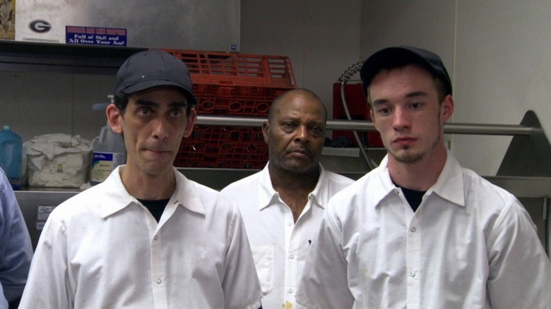Three chefs looking sombre wearing white shirts inside kitchen