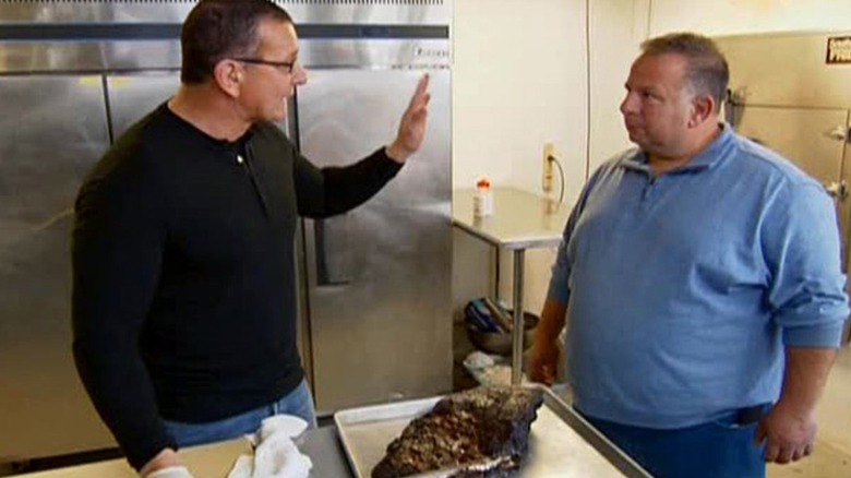 Robert Irvine gestures at chef in front of cooked meat in kitchen